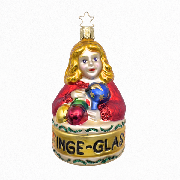 Decorative glass Christmas ornament of a blonde girl holding several small glass ornaments with the caption "INGE-GLAS" underneath her and a gold star top for hanging by Inge Glas of Germany.