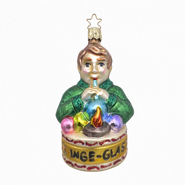 A glass ornament depicting a stylized male character in green, seated at a table with a pipe blowing glass, surrounded by colorful decorative elements and a sign at the bottom that says "INGE-GLAS" on a gold star top for hanging.