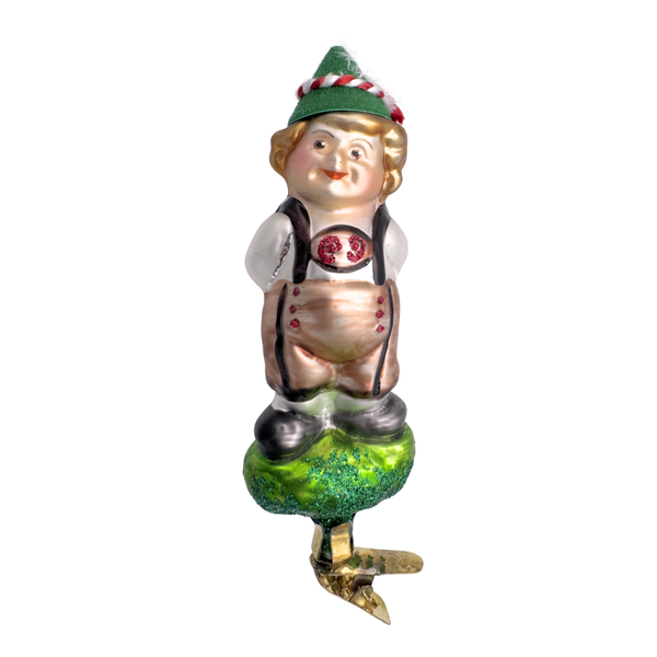 A clip on ornament featuring a glass figurine of a Tirolian Tot, with traditional clothing and a green hat, standing on a green base with a gold clip for hanging.