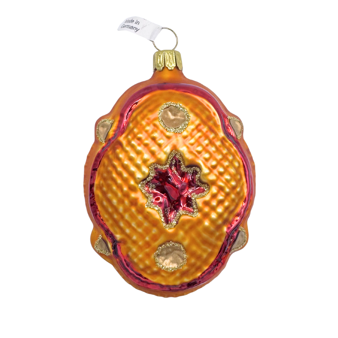 Decorative glass ornament in the shape of a Victorian pillow in shades of warm amber and burgundy with scalloped edges and a star design in the center on a white background