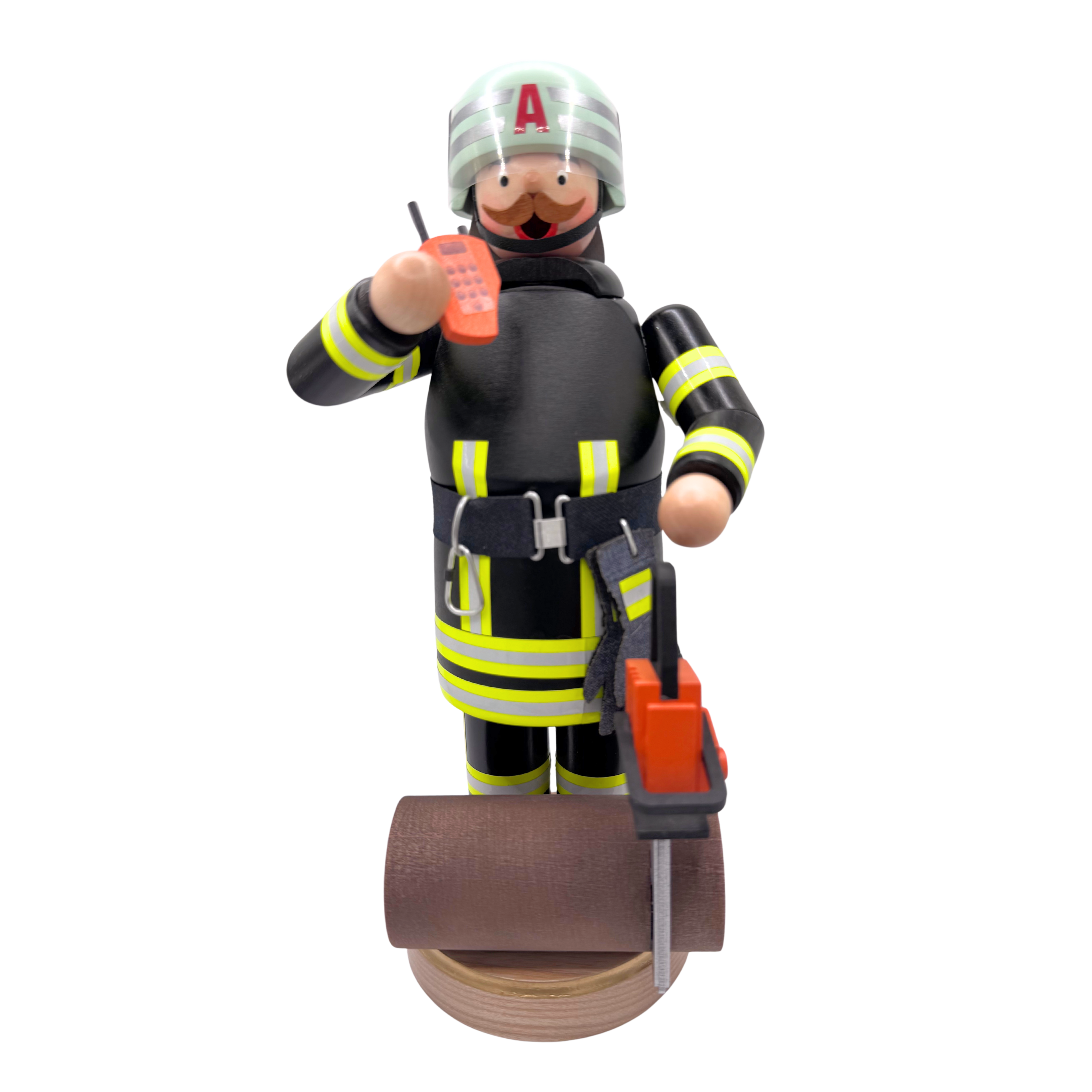 Wood figure of a firefighter dressed in black and fluorescent yellow holding a phone with log and chainsaw sitting on the base in front of his feet, on a white background